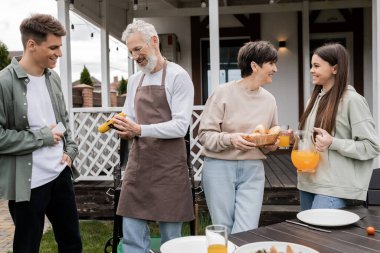 parents day, tattooed father holding tongs with grilled corn near son with smartphone, mother smiling and talking with teenage daughter, bbq party, translation of tattoo: harm none do what you will  clipart
