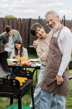 parents day, middle aged couple smiling during bbq party, bearded man holding tongs near bbq grill and wife, daughter and son on blurred background, translation of tattoo: harm none do what you will  clipart