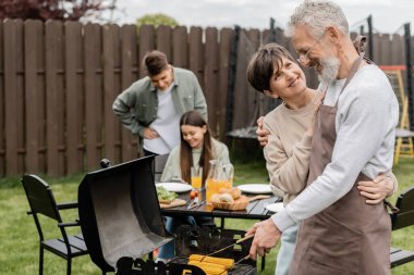 parents day, middle aged couple smiling during family bbq party, bearded tattooed man holding tongs near bbq grill, grilling corn, daughter and son, translation of tattoo: harm none do what you will  clipart