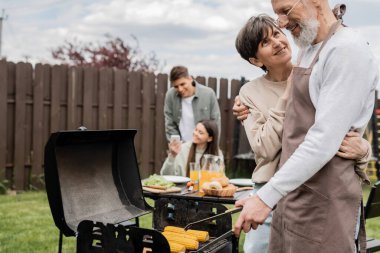parents day, middle aged couple hugging during family bbq party, bearded tattooed man holding tongs near bbq grill, grilling corn, daughter and son, translation of tattoo: harm none do what you will  clipart