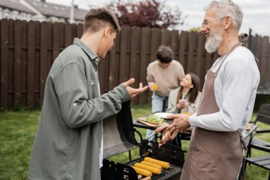 tattooed and bearded father and son preparing food on bbq grill, grilling corn, communication, family bbq party, middle aged, happy parents day, translation of tattoo: harm none do what you will  clipart