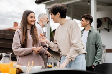 Mutlu orta yaşlı bir anne elinde vişneli domates tutarken ve genç kız ile konuşurken, aile mangal partisi, arka planda baba ve oğul, mutlu aile günü konsepti, yaz mevsimi,