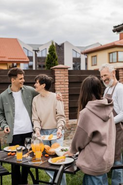 family grill party, celebration of parents day, happy young adult son hugging middle aged mother, bearded father in apron looking at teenage daughter, translation of tattoo: harm none do what you will  clipart