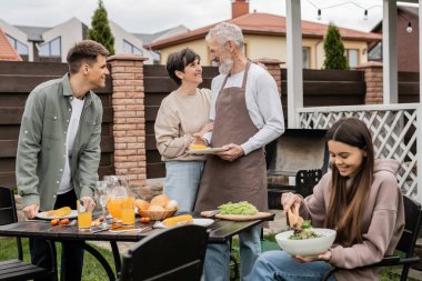 middle aged couple looking at each other during bbq party, young adult son looking at cheerful parents, teenage girl mixing salad, parents day, translation of tattoo: harm none do what you will clipart