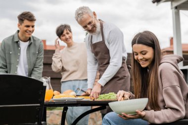 Neşeli genç kız salatayı neşeli orta yaşlı anne, baba ve yetişkin erkek kardeşin yanında karıştırıyor aile bbq partisi, ebeveynler gününü kutluyor, pozitiflik, yaz zamanı, aile evinin arka bahçesi. 