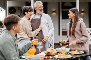 celebrating parents day, happy middle aged couple looking at joyful teenage girl serving salad near adult brother with glass of orange juice, love, family grill party, summer  clipart