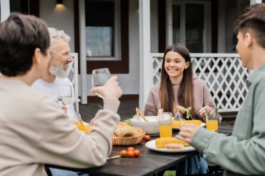 Mutlu genç kız aile kutlaması sırasında neşeli ebeveynlere bakıyor, yazlığın arka bahçesinde oturuyor, birlikte vakit geçiriyor, ızgara bbq yemeği yiyor, mutlu ebeveynler günü konsepti. 