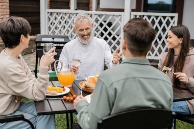 Aile partisinde yetişkin oğluna bakan neşeli orta yaşlı bir adam, yazlığın arka bahçesinde oturuyor, birlikte vakit geçiriyor, ızgara bbq yemeği yiyor, mutlu ebeveynler günü konsepti. 