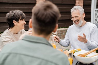 cheerful middle aged man looking wife near adult son and gesturing during bbq party, blurred foreground, backyard of summer house, spending time together, eating grilled bbq food, parents day concept  clipart