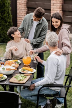 Positive siblings hugging and talking to middle aged parents with wine near summer food during barbeque party and parents day celebration at backyard, family love and unity concept clipart