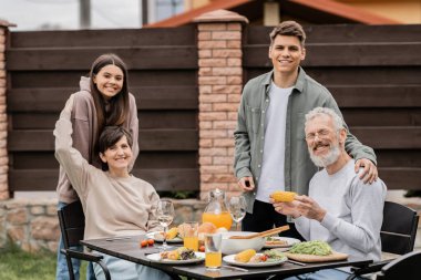 Çocukların yanında kameraya bakan orta yaşlı ebeveynler ve aile barbeküsü sırasında lezzetli yaz yemekleri ve Haziran 'da arka bahçede aileler günü kutlaması, aile sevgisi ve birlik kavramı.
