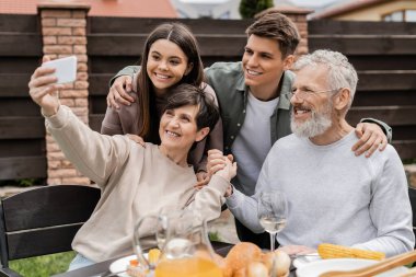 Gülümseyen orta yaşlı bir kadın kocasının elini tutarken ailesi ve çocuklarıyla telefonda selfie çekiyor. Ve arka bahçede veli günü kutlaması, mutlu aile günü konsepti.
