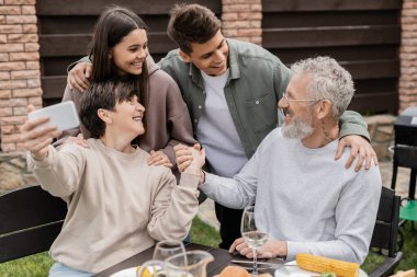 Çocukların konuştuğu neşeli bir aile yaz yemeği sırasında akıllı telefondan selfie çekerken ve aile günü kutlaması Haziran 'da arka bahçede, mutlu aile günü konsepti.