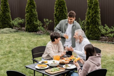 Heyecanlı ve neşeli orta yaşlı ebeveynler arka bahçedeki BBS partisinde çocukların yanında şarap kadehleriyle ve yaz yemekleriyle kadeh kaldırıyorlar aile bağları kavramına değer veriyorlar, birlikte vakit geçiriyorlar.