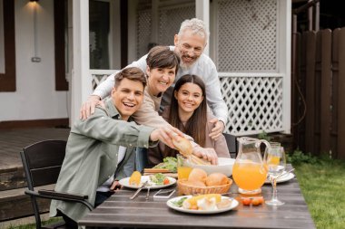 Cheerful middle aged parents looking at camera while hugging kids near tasty summer food and parents day celebration at backyard in june, special day for parents concept, traditions and celebrations clipart