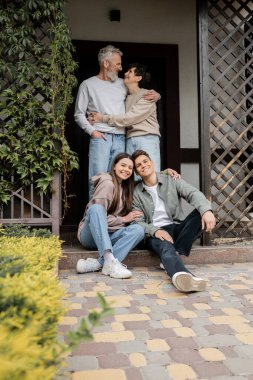 Olumlu genç bir kız, küçük kardeşine sarılıyor ve orta yaşlı ebeveynlerin aile günü kutlamaları, aile gelenekleri ve kutlama konsepti sırasında evinin verandasında otururken kameraya bakıyor.