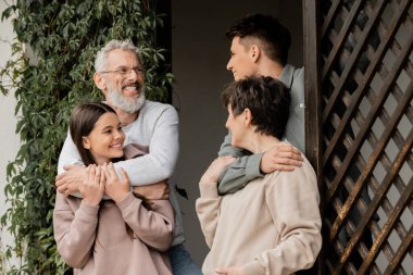 Positive young man hugging middle aged mother and talking to father and teenage sister during parents day celebration while standing on porch of house, parent-child relationship concept clipart