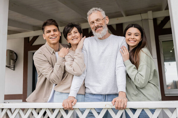 parents day celebration, middle aged parents hugging with cheerful daughter and son on porch of summer house, looking at camera, family reunion, bonding, modern parenting, moments to remember 