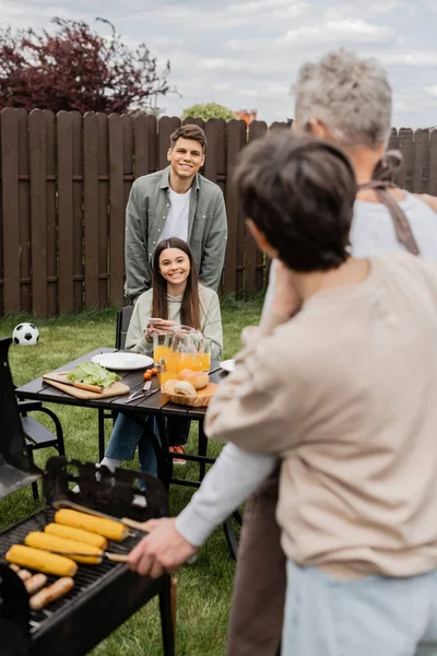 両親の日のお祝いお父さんとお母さんを見ている幸せな兄弟背景 家族のBbq グリルパーティー スマートフォンを使用して グリルBbqで食べ物を準備し 現代的な子育て — ストック写真
