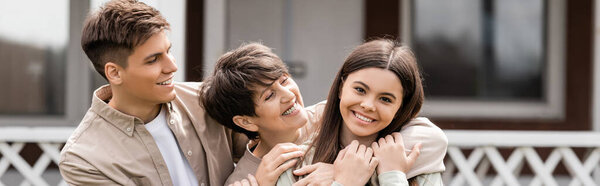 happy parents day, mother hugging cheerful teenage daughter near young adult son on backyard of family house, celebration, bonding, moments to remember, modern parenting, banner 