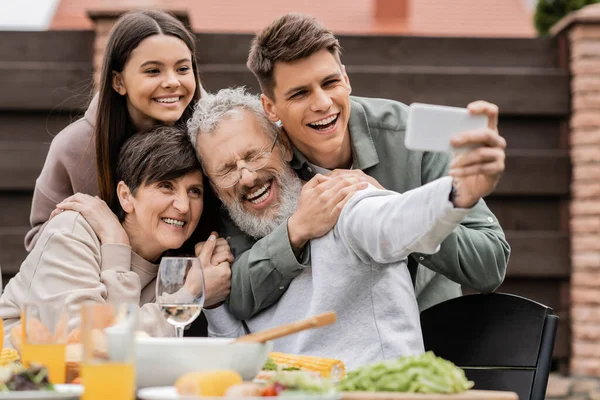Cheerful kids hugging middle aged parents while taking selfie on smartphone together during barbeque party and parents day celebration at backyard in june, happy parents day concept
