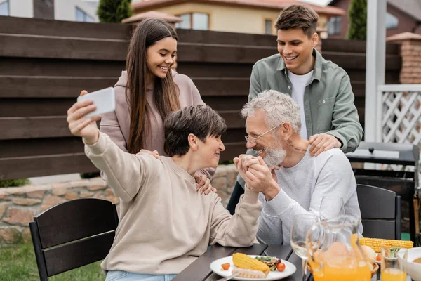 Gülümseyen orta yaşlı bir kadın kocasının elini tutuyor ve yaz partisinde çocuklarla birlikte akıllı telefondan selfie çekiyor. Arka bahçede aileler günü kutlaması, mutlu aileler günü konsepti.