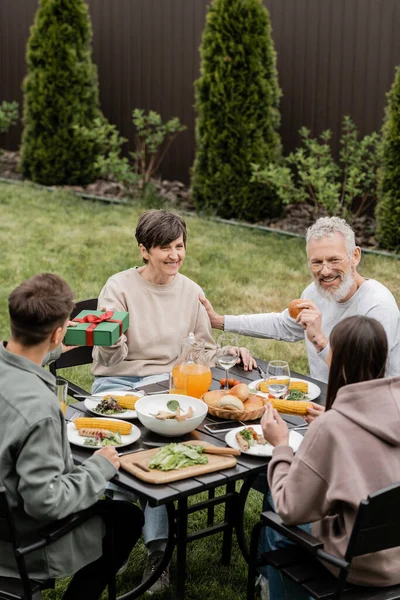 stock image Young son giving gift box to cheerful middle aged mother near sister and father while celebrating parents day and family bbq party at backyard in june, cherishing family bonds concept