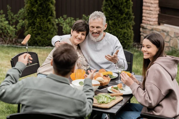 Smiling middle aged parents hugging and talking to children near summer food during bbq party and parents day celebration at backyard in june, cherishing family bonds concept, special occasion