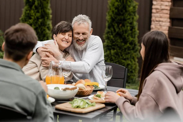Cheerful middle aged couple hugging near blurred kids and summer food during bbq party and parents day celebration at backyard in june, special day for parents concept, special occasion