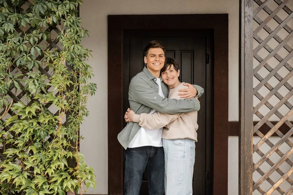 stock image Cheerful young man hugging middle aged mother and looking at camera while standing together on porch of house during parents day celebration, quality time with parents concept 