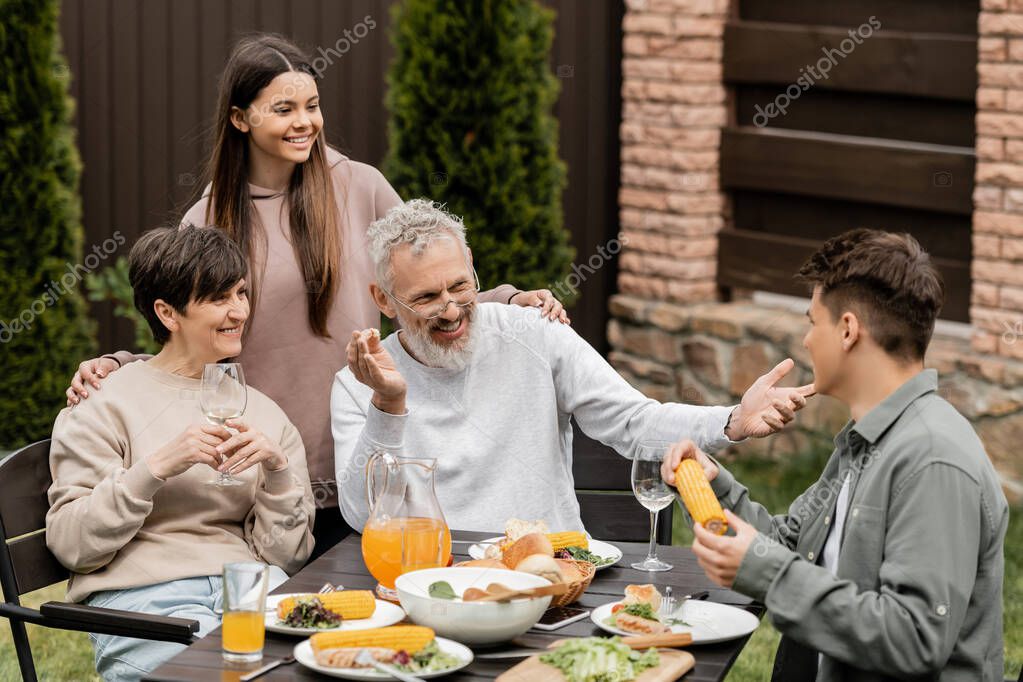 Cheerful middle aged man talking to young soon while sitting near family and summer food during bbq party and parents day celebration at backyard, special day for parents concept, tradition and celebration