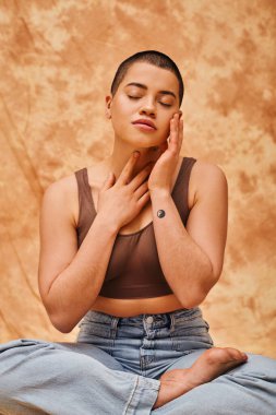 body image, relaxation, curvy young and tattooed woman in jeans and crop top sitting with crossed legs on mottled beige background, closed eyes, personal style, self-acceptance, generation z  clipart