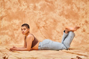 body positivity, casual attire, curvy young and tattooed woman in jeans and crop top lying on mottled beige background, looing at camera, denim fashion, personal style, self-acceptance, generation z  clipart