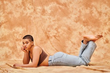 body positivity, representation of body, curvy young and tattooed woman in jeans and crop top lying on mottled beige background, looing at camera, denim fashion, personal style, generation z  clipart