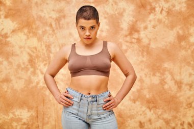 body positivity movement, curvy young woman with tattoos posing in jeans and crop top on mottled beige background, hands on hips, representation of body, confidence, casual attire, generation z  clipart