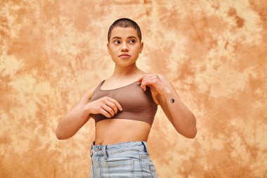 body positivity movement, curvy young woman with tattoos posing in jeans and crop top on mottled beige background, representation of body, confidence, casual attire, generation z, looking away clipart
