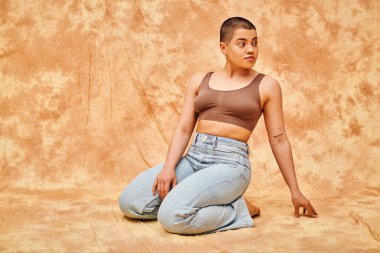 body positivity, denim fashion, curvy and tattooed woman in jeans and crop top sitting on mottled beige background, casual attire, looking away, self-acceptance, generation z, body diversity  clipart
