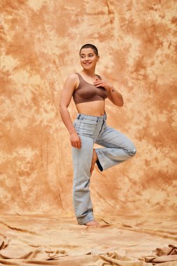 body positivity, confident, curvy and happy woman in crop top and jeans posing on mottled beige background, casual attire, self-acceptance, generation z, tattooed, smile, full length, denim fashion  clipart