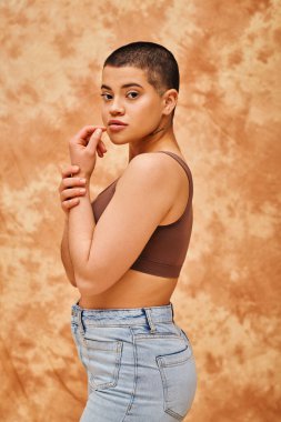 representation of body, curvy and young woman in crop top and jeans posing on mottled beige background, short haired, self-acceptance, generation z, tattooed, different shapes, looking at camera clipart