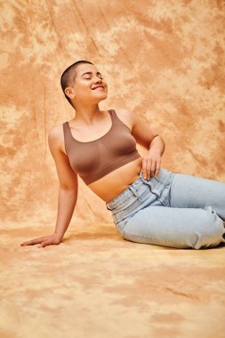 body positivity, denim fashion, curvy and pleased woman in jeans and crop top sitting on mottled beige background, tattooed, casual attire, self-acceptance, generation z, body diversity  clipart