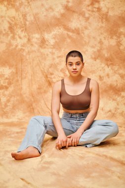 body positivity, denim fashion, curvy tattooed woman in jeans and crop top sitting on mottled beige background, casual attire, looking at camera, self-acceptance, generation z, short haired model  clipart