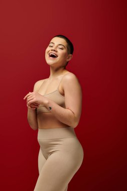 Kendini kabullenme, kırmızı arka planda bej iç çamaşırlı, etkileyici ve dövmeli kadın, vücut pozitifliği, kıvrımlı moda, kıvrımlı model, Z nesli, kendini beğenmiş, kısa saçlı. 