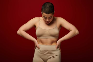 body positive, self-esteem, young woman with short hair and tattoo posing with hands on hips on burgundy background, dark red, curvy fashion, comfortable in skin, female underwear, fashion model  clipart