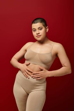 Vücut pozitifliği, kendine güven, kısa saçlı ve dövmeli genç kadın burgundy arka planda poz veriyor, koyu kırmızı, kıvrımlı model, kadın iç çamaşırı, moda, vücut tipi 