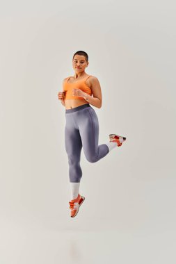 Vücut pozitifliği, kısa saçlı genç bir kadının gri arka planda zıplaması, kıvrımlı moda, kadın fitness, güçlendirme, motivasyon, spor kıyafeti, kuvvet ve sağlık, vücut imajı. 
