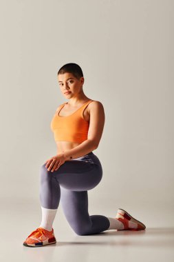Vücut pozitifliği hareketi, gri arka planda dizlerinin üzerinde duran kısa saçlı genç bir kadın spor giyim, güçlendirme, motivasyon, yükseltilmiş eller, güç ve sağlıkla çalışma. 