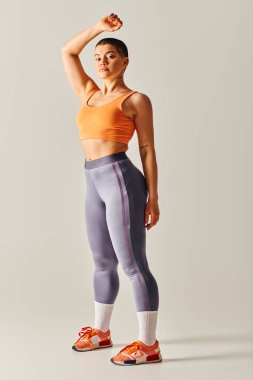 Vücut özgüveni, atletik ve kısa saçlı kadın gri arka planda poz veriyor, kıvrımlı fitness modeli, kaldırılmış elle duruyor, dayanıklılık ve güç, Z nesli, tam boy 