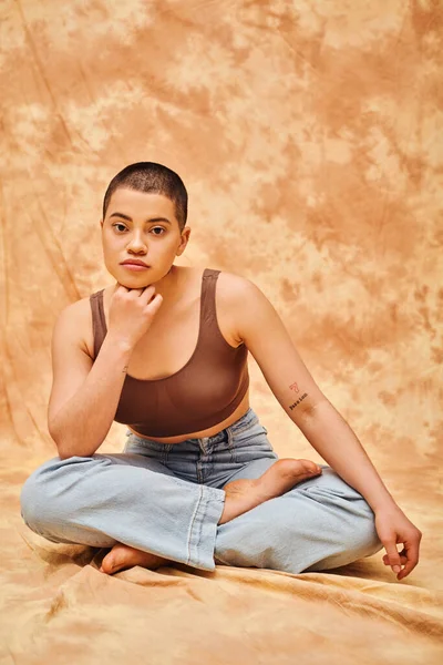 body confidence, acceptance, curvy young and tattooed woman in jeans and crop top sitting with crossed legs on mottled beige background, personal style, self-acceptance, generation z, denim fashion