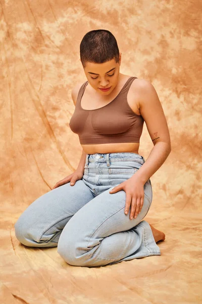 body positivity, denim fashion, curvy and tattooed woman in jeans and crop top sitting on mottled beige background, personal style, self-acceptance, generation z, casual attire