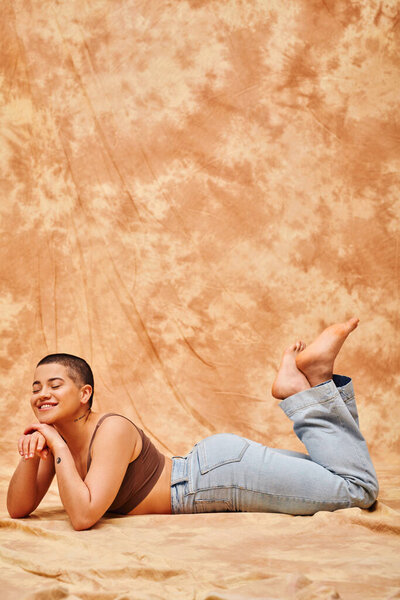 body positivity, representation of body, curvy and tattooed woman in jeans and crop top lying on mottled beige background, smiling with closed eyes, denim fashion, personal style, generation z 
